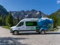Biały bus Tatrzańskiego Parku Narodowego z grafiką górskiego krajobrazu, przypominającego Morskie Oko, stoi na kamiennej drodze.