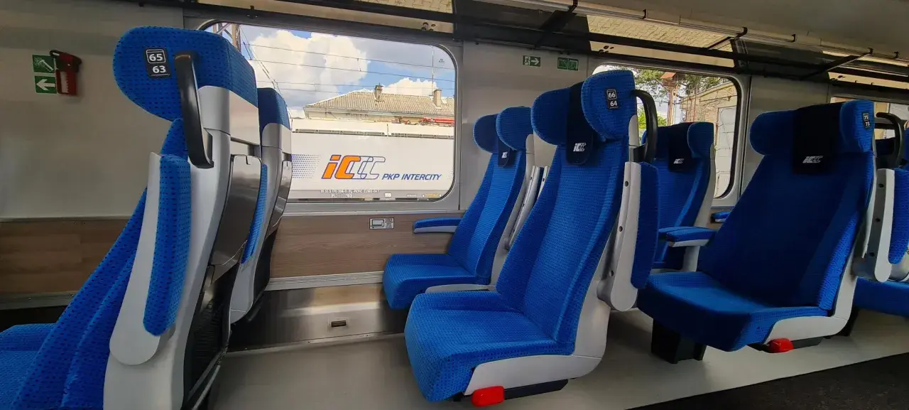 Wygodne, niebieskie fotele w wagonach PKP Intercity bez przedziałów. Widok na mijany krajobraz i inne wagony.