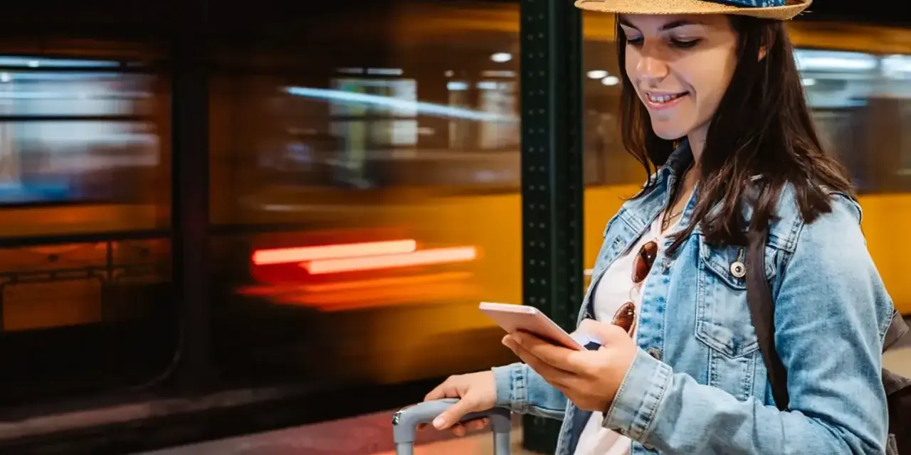Kobieta z walizką na peronie sprawdza telefon, szukając informacji, jak kupić bilet na pociąg.