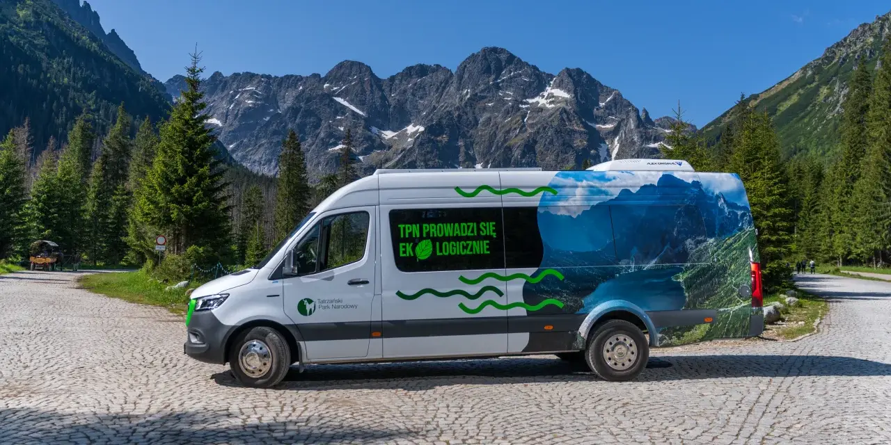Biały bus Tatrzańskiego Parku Narodowego z grafiką górskiego krajobrazu, przypominającego Morskie Oko, stoi na kamiennej drodze.
