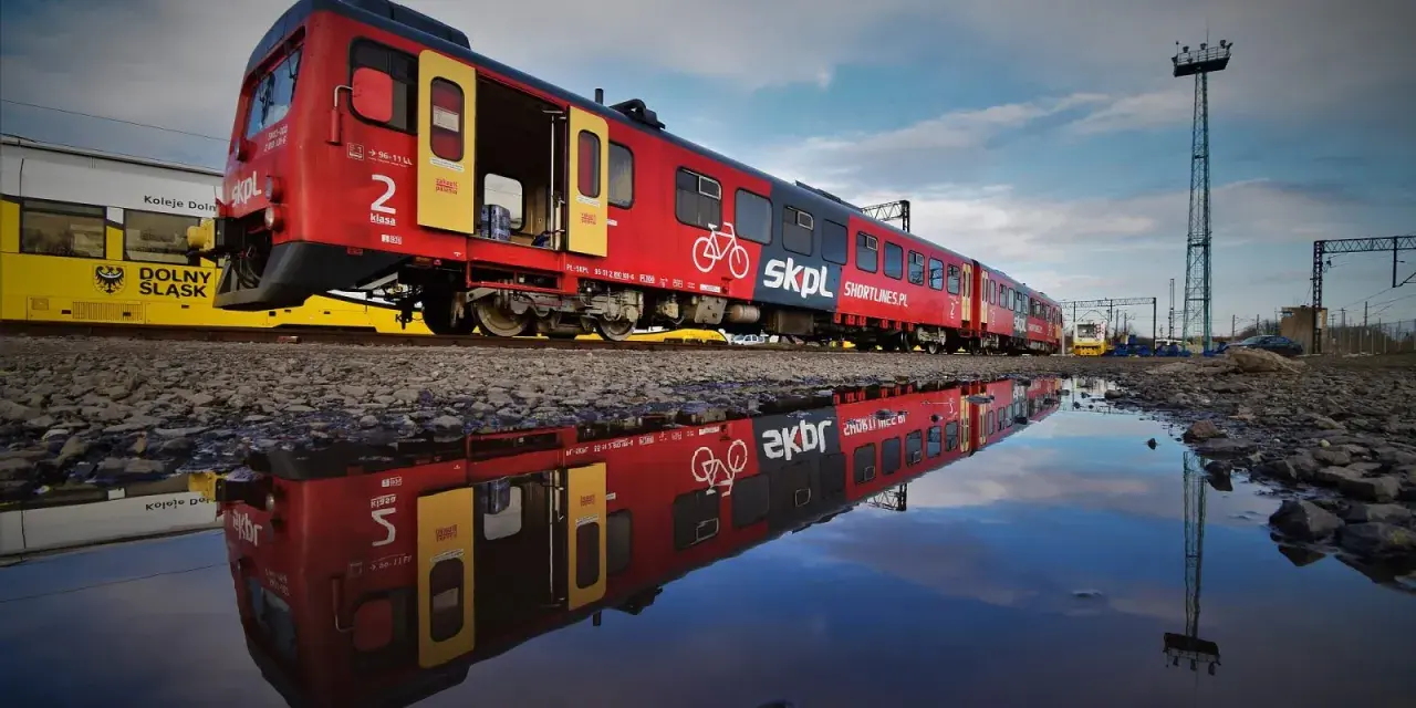 Czerwony pociąg SKPL, stowarzyszenie kolejowych przewozów lokalnych, odbija się w kałuży. Obok żółty tramwaj Kolei Dolnośląskich.