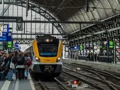Grupa ludzi czeka na peronie na żółto-szary pociąg. Znaki informacyjne wskazują, jak dojechać PKP.