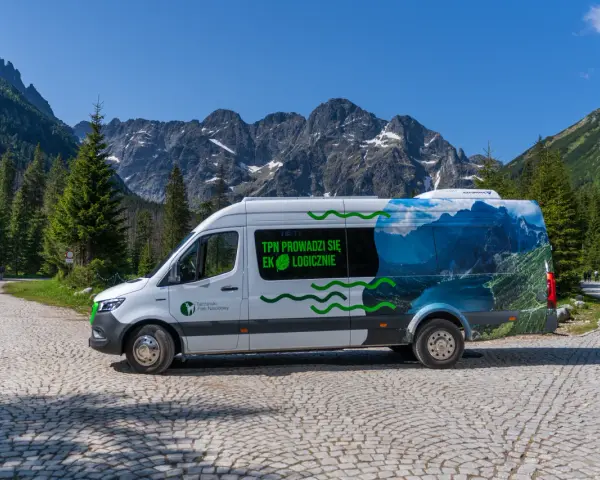 Biały bus Tatrzańskiego Parku Narodowego z grafiką górskiego krajobrazu, przypominającego Morskie Oko, stoi na kamiennej drodze.