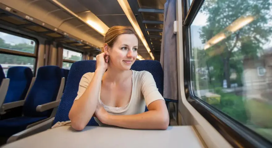 Uśmiechnięta kobieta podróżuje pociągiem z Krakowa do Wiednia, patrząc przez okno na mijane krajobrazy.