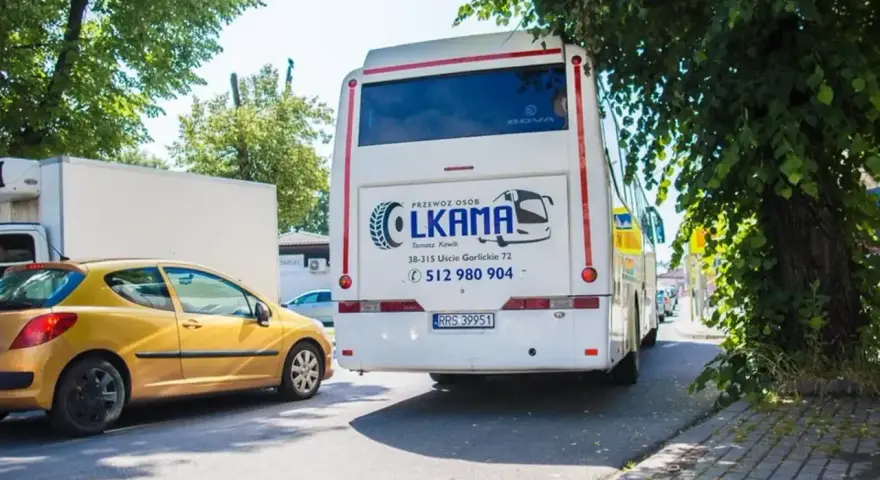 Autobus LKAMA z napisem "Voyager Nowy Sącz Gorlice" stoi obok żółtego samochodu.