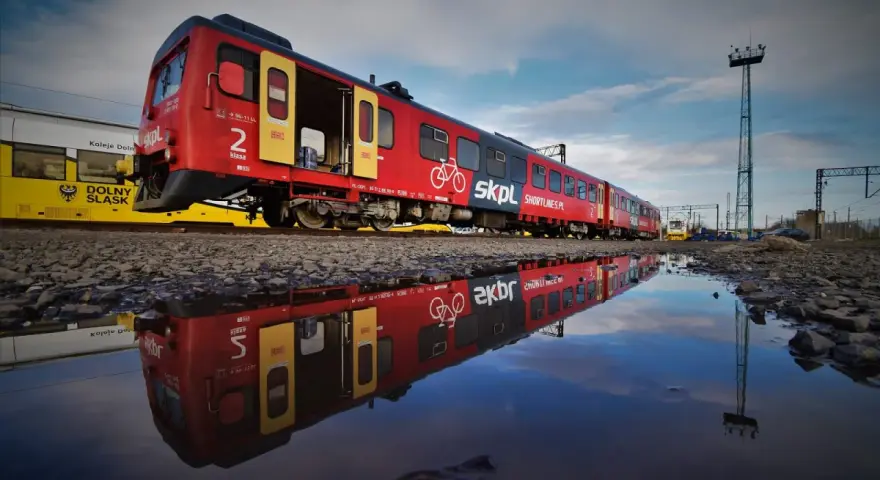 Czerwony pociąg SKPL, stowarzyszenie kolejowych przewozów lokalnych, odbija się w kałuży. Obok żółty tramwaj Kolei Dolnośląskich.