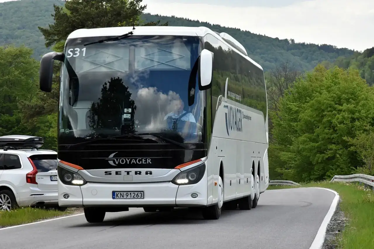 Autokar Voyager na trasie Nowy Sącz - Gorlice, z kierowcą za kierownicą, jedzie po krętej drodze wśród zielonych wzgórz.
