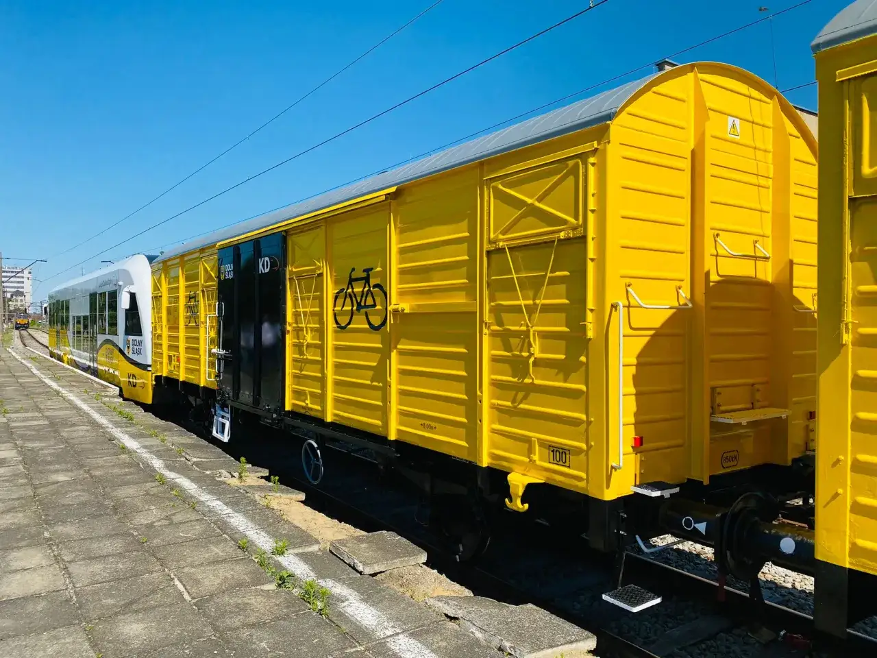 Żółty wagon towarowy z symbolem roweru, część pociągu Koleje Dolnośląskie, stoi na bocznicy.
