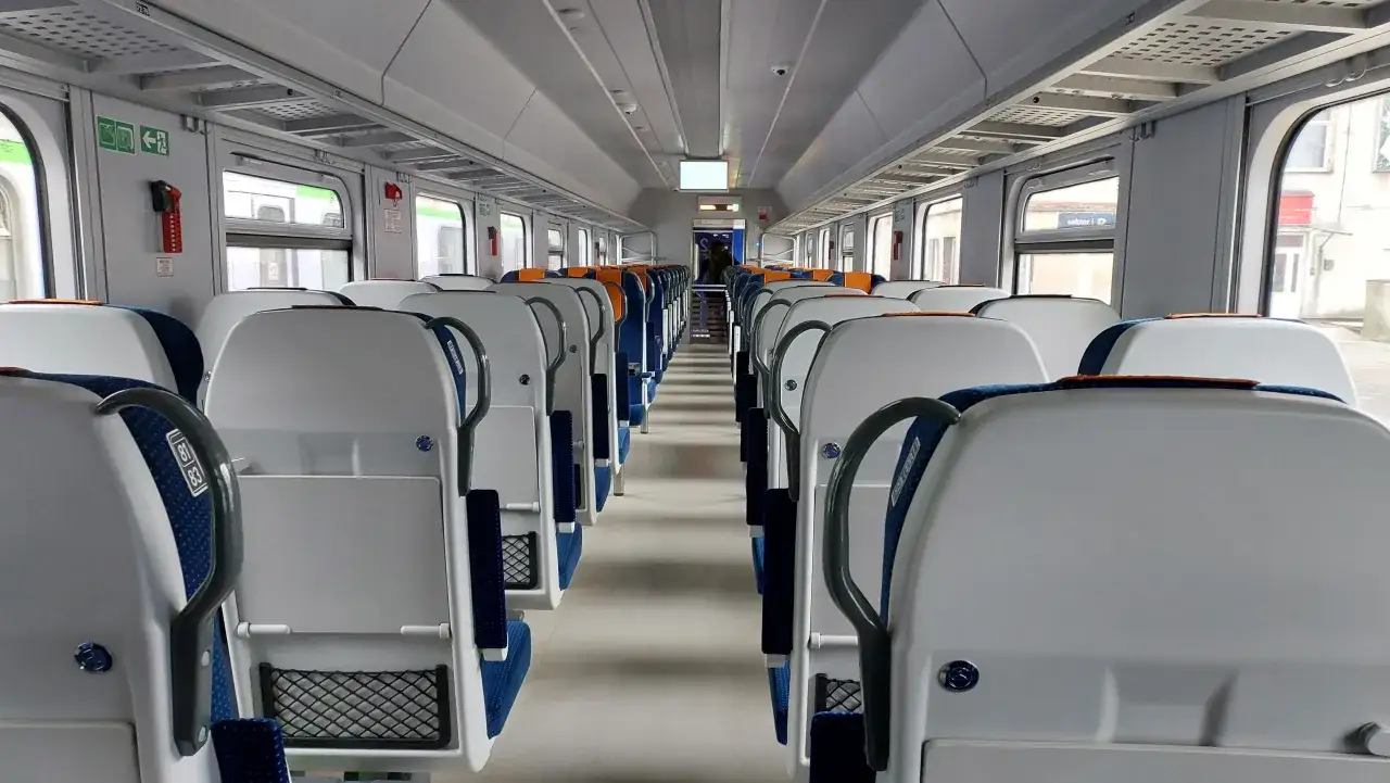 Wnętrze nowoczesnego pociągu z rzędami białych siedzeń i niebieskimi akcentami. Intercity wagony bez przedziałów oferują przestronną podróż.