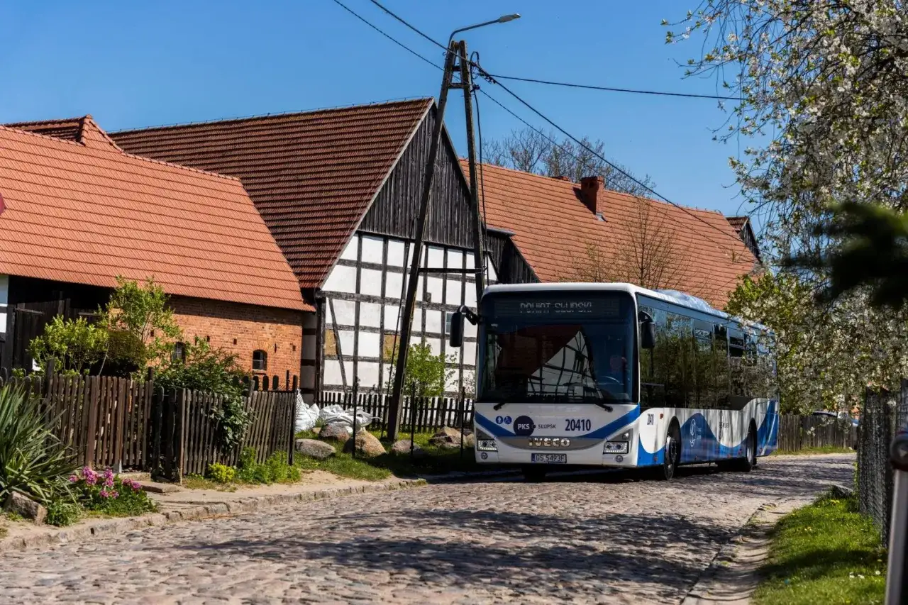 Autobus PKS Słupsk jedzie brukowaną ulicą w Jarosławcu, mijając tradycyjne domy z czerwonymi dachami.