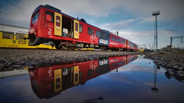 Czerwony pociąg SKPL, stowarzyszenie kolejowych przewozów lokalnych, odbija się w kałuży. Obok żółty tramwaj Kolei Dolnośląskich.