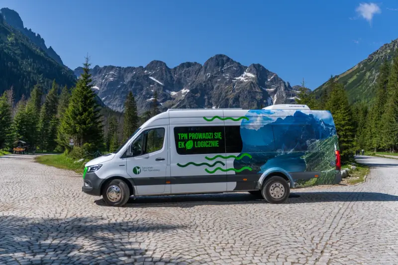 Biały bus Tatrzańskiego Parku Narodowego z grafiką górskiego krajobrazu, przypominającego Morskie Oko, stoi na kamiennej drodze.
